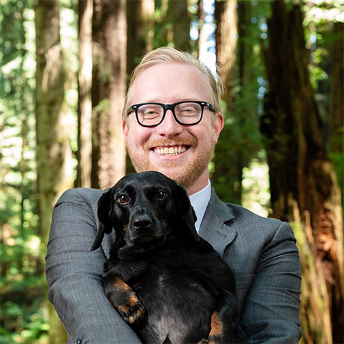 A well dressed man stands in the forest and holds a dog while smiling