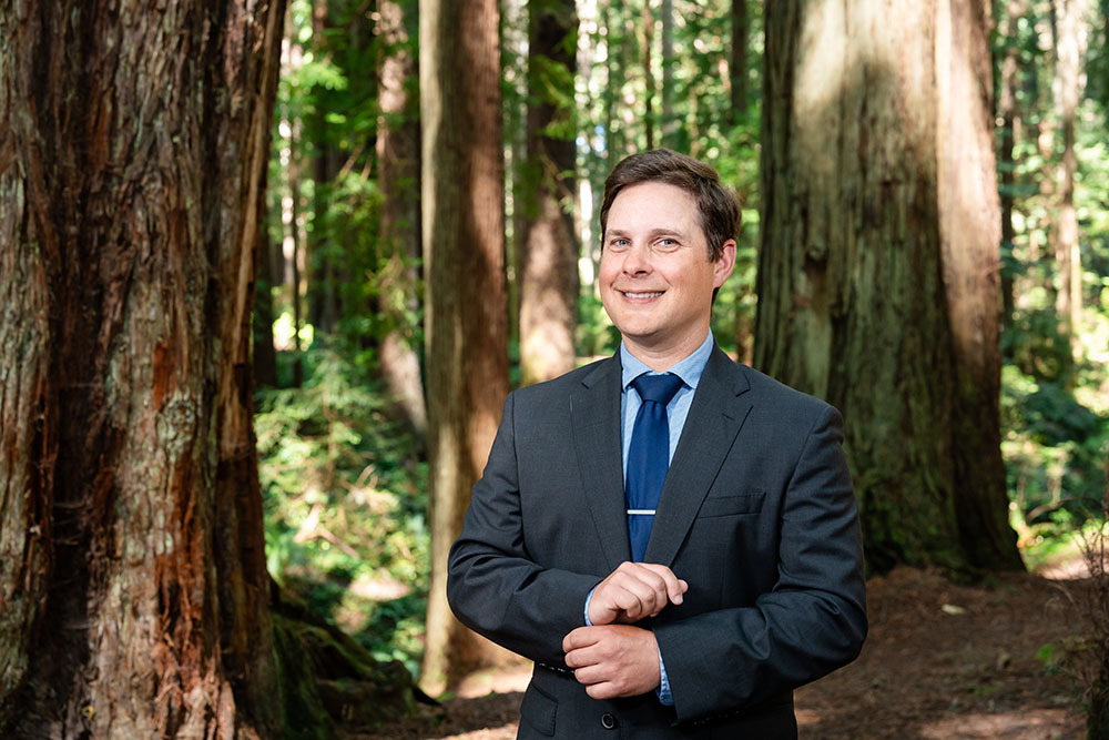 A well dressed man stands in the forest and smiles