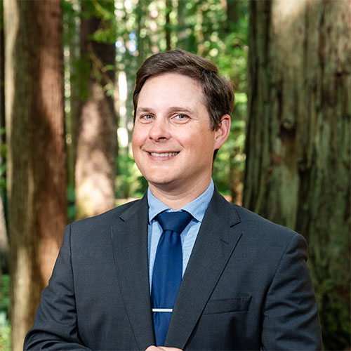 A well dressed man stands in the forest and smiles
