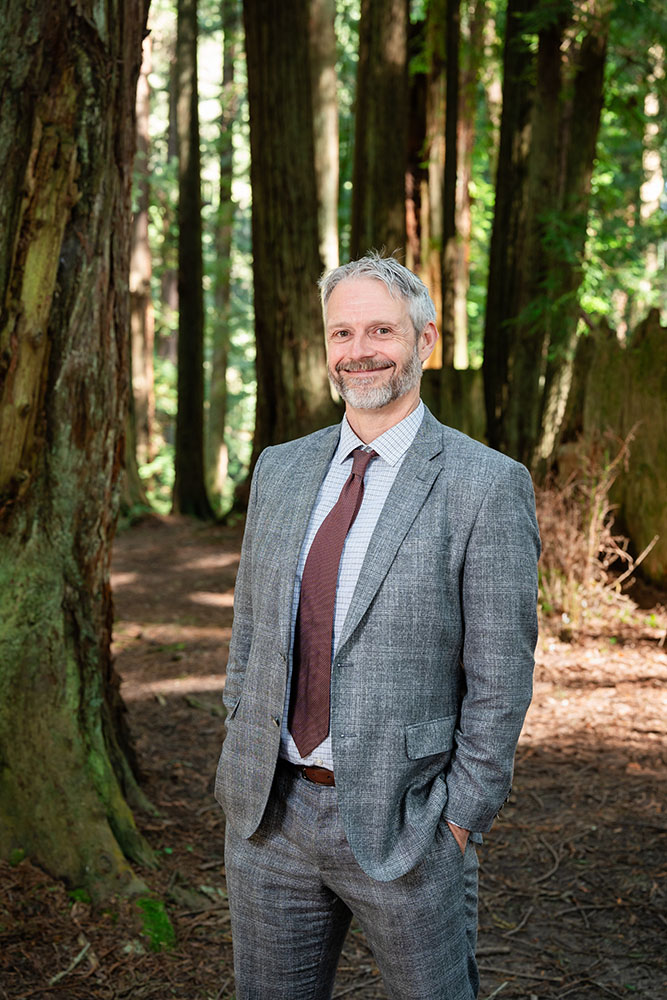 A well dressed man stands in the forest and smiles