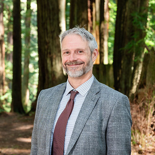 A well dressed man stands in the forest and smiles